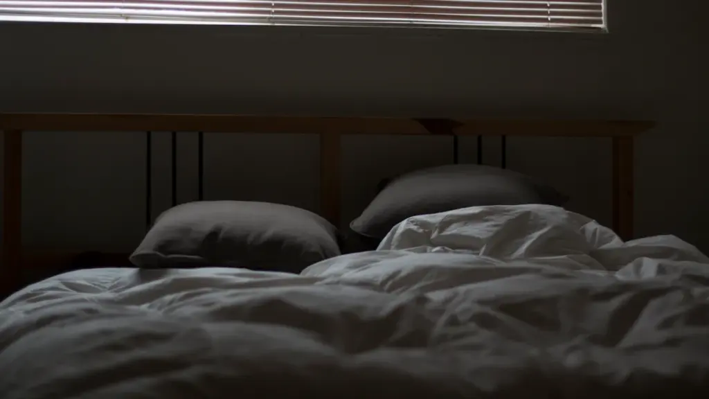 Ein ungemachtes Bett mit heruntergelassenen Jalousien bei abgedunkeltem, warmem Tageslicht im Schlafzimmer.