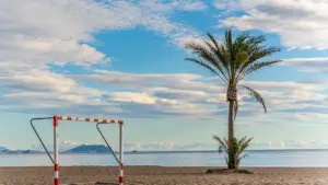 Ein Fußballtor steht einsam am weiten Sandstrand unter blauem Himmel.