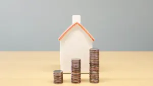 Ein kleines Hausmodell neben Münzstapeln auf einem Schreibtisch zur Visualisierung von Immobilienfinanzierungen.