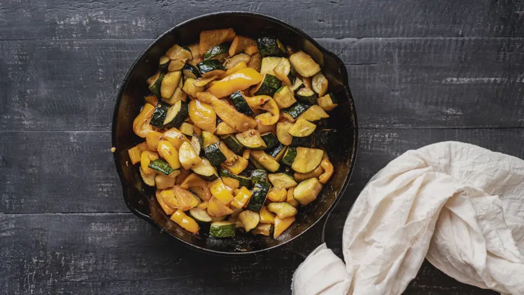 Eine gusseiserne Pfanne mit gebratenen Zucchini- und Paprikastücken auf dunklem Holzuntergrund.