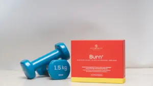 Blaue Hanteln und eine Nahrungsergänzungsmittel-Packung auf einem hellen Hintergrund.