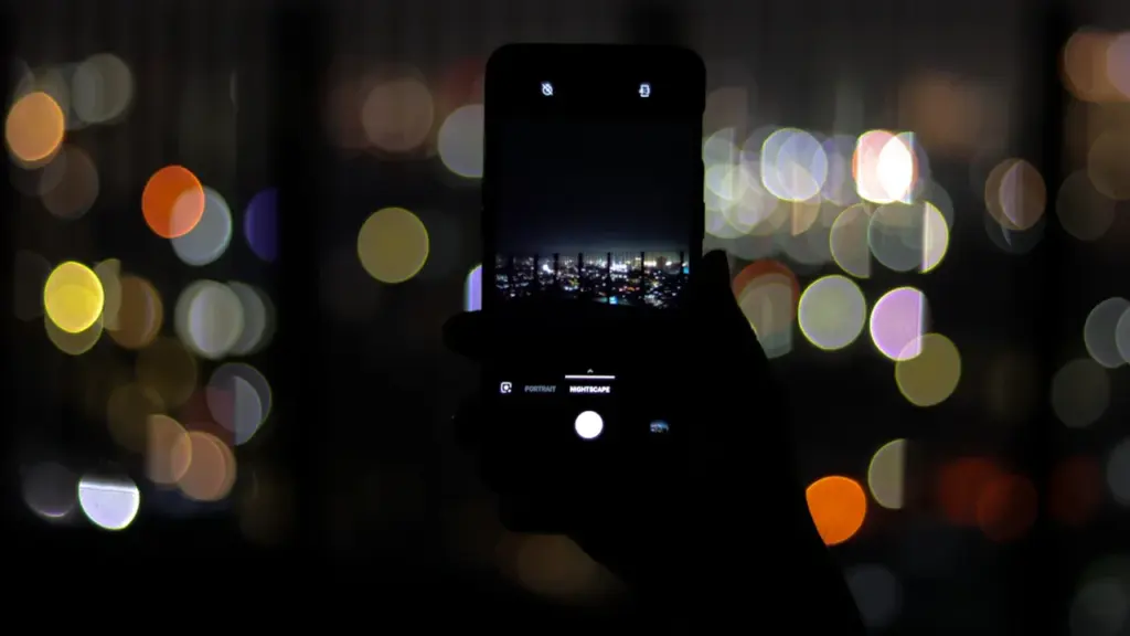 Eine Hand hält ein Smartphone, das eine nächtliche Stadtlandschaft vor unscharfen bunten Lichtern fotografiert.
