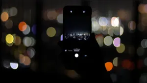 Eine Hand hält ein Smartphone, das eine nächtliche Stadtlandschaft vor unscharfen bunten Lichtern fotografiert.