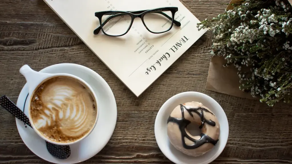 Eine Brille liegt auf einem Notizblock neben Kaffee und einem Krapfen auf Holztisch.