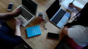 Zwei Personen arbeiten an Laptops an einem gemeinsamen Holztisch in einem modernen Büro.
