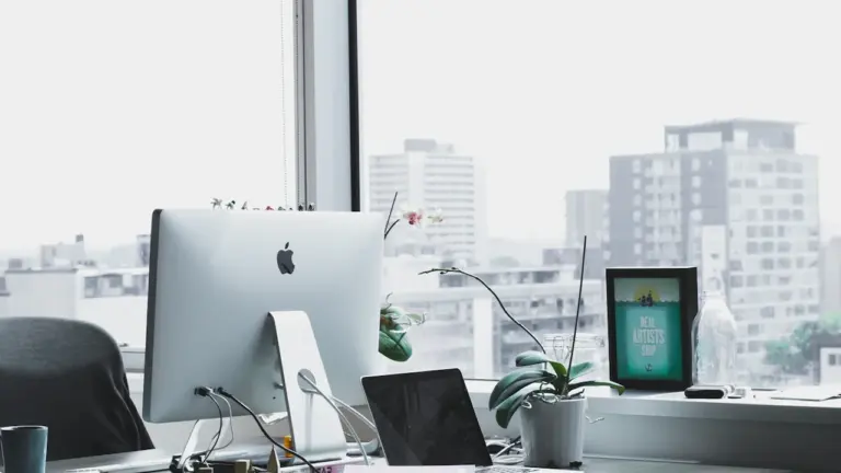 Moderner Schreibtisch mit iMac vor einem Fenster in einem hellen Bürogebäude.