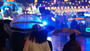 Besucher nutzen bei regnerischem Wetter Regenschirme auf dem beleuchteten Münchner Oktoberfest Gelände.