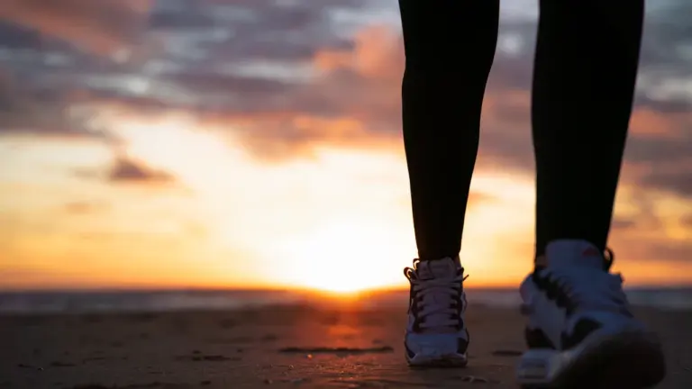 Eine Person geht in Sportschuhen am Strand in den Sonnenuntergang.