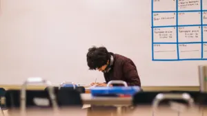 Ein konzentrierter Student schreibt an seinem Schreibtisch vor einer großen, beschrifteten Lehrtafel.