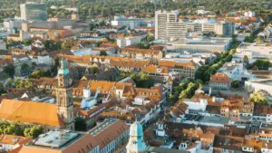 Luftaufnahme einer Stadt mit roten Ziegeldächern, einer prominenten Kirchturmspitze und modernen Bürogebäuden im Hintergrund.